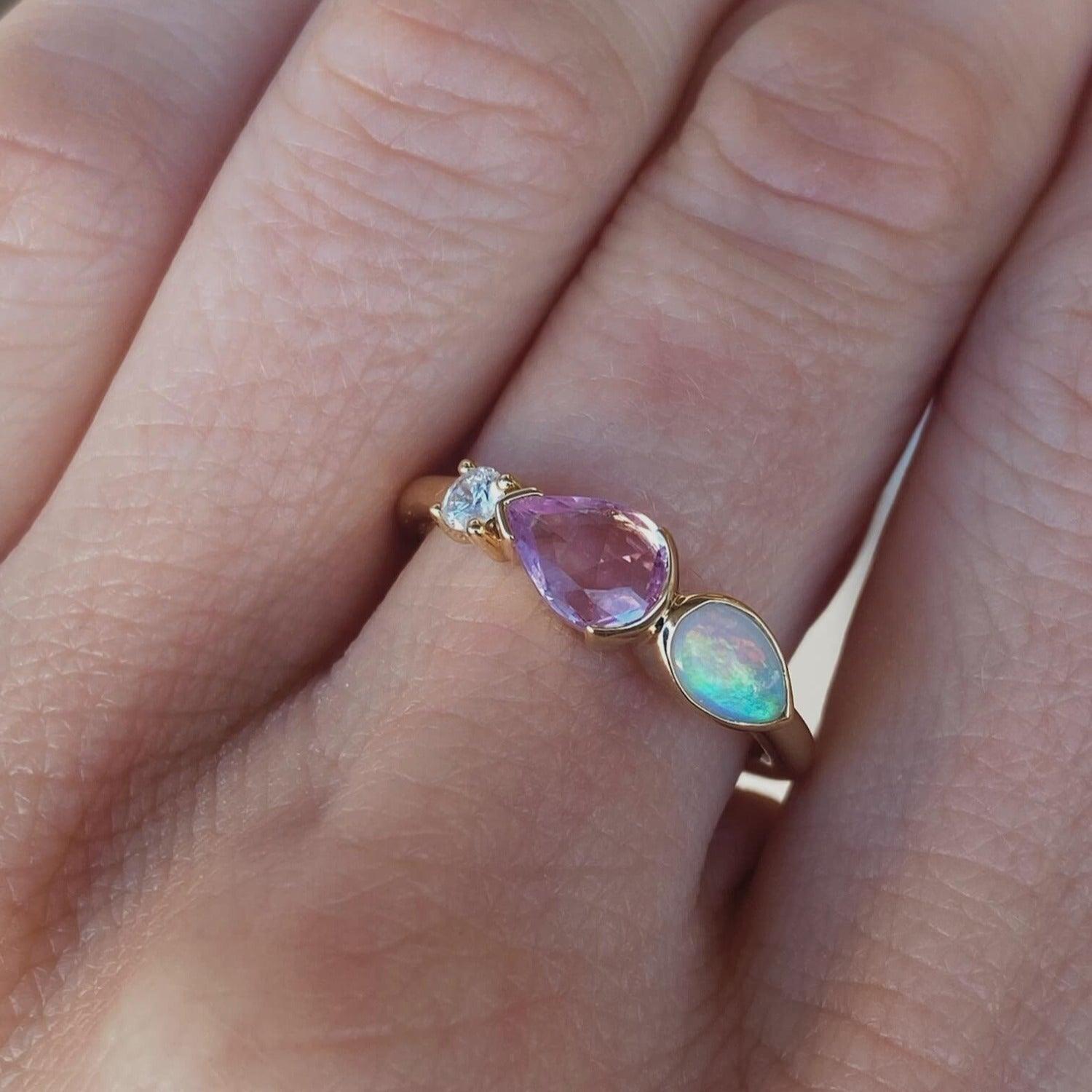 Image of a linear band on a hand. This ring is in 14K yellow gold and has a rose cut pink sapphire pear, opal pear, and small white diamond round.