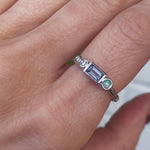 Image of a marrow fine linear band on a pointer finger. The one of a kind ring has diamonds, a spinel baguette, and a small opal round. All stones are bezel set and in 14K white gold.