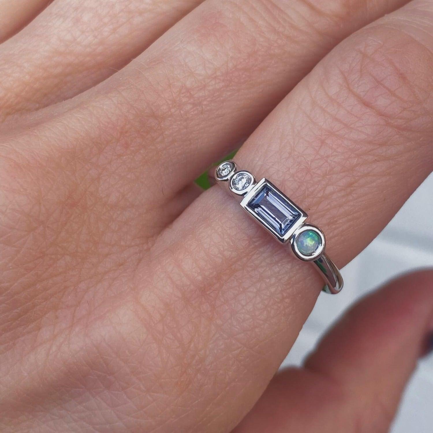 Image of a marrow fine linear band on a pointer finger. The one of a kind ring has diamonds, a spinel baguette, and a small opal round. All stones are bezel set and in 14K white gold.