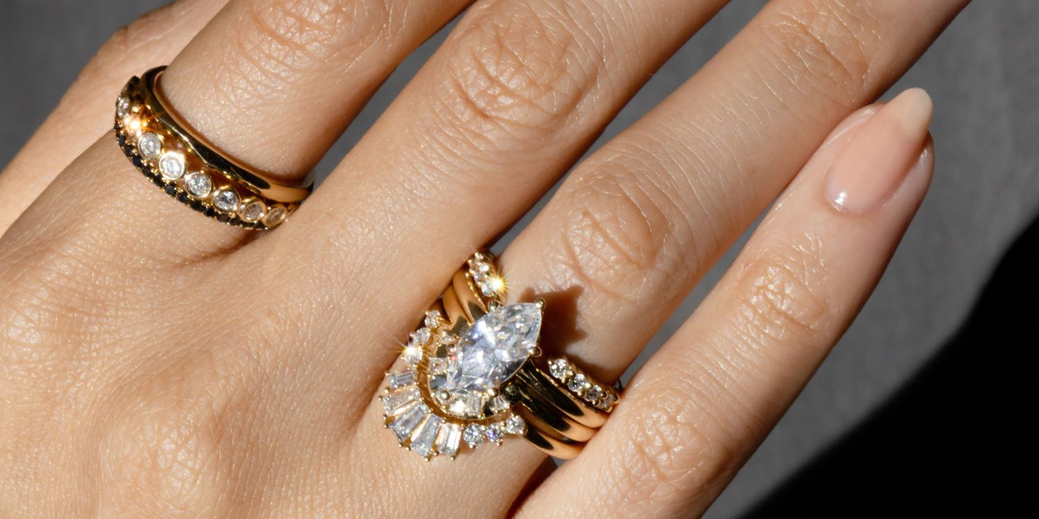 CLose up of hand featuring a marquise diamond engagement ring, stacked with multiple Marrow Fine rings