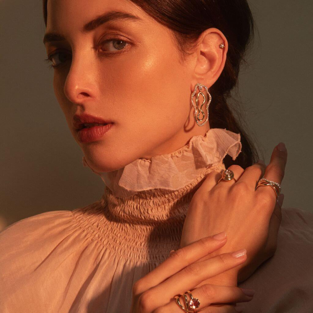 Close up of woman looking at the camera with hands positioned on top of each other, laying on her left clavicle and wearing Marrow Fine jewelry.